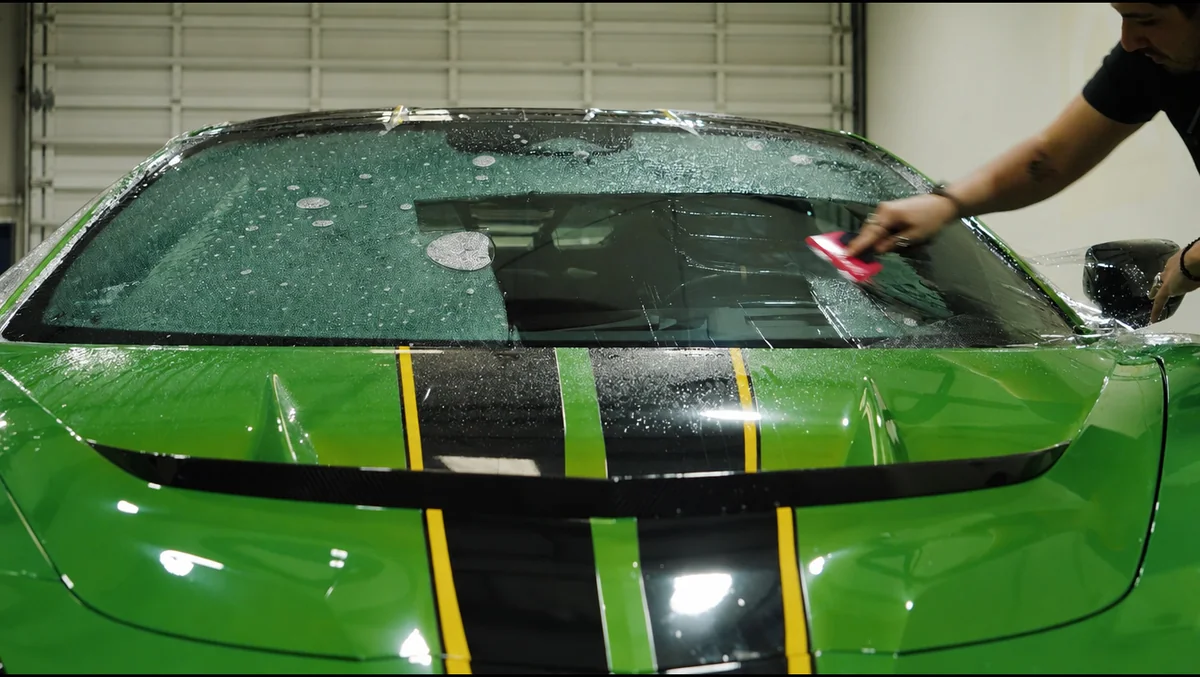 An Auto Trim Plus installer squeegeeing clear protection film onto the windshield of a green sports car